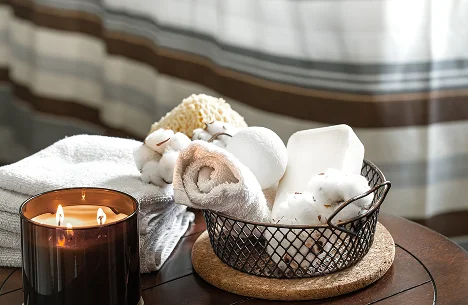 Przytulna scena spa z zapaloną świecą, zwiniętymi ręcznikami i przyborami do kąpieli w koszu na drewnianym stole. Miękkie światło tworzy relaksującą atmosferę.